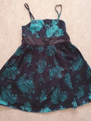 Klänning - Snygg klänning med grönt blommönster och tunna axelband. Klänningen har en markerad midja med ett brett band och lätt utställd kjol. Perfekt för dig som gillar en feminin och trendig stil. Finns röd fläck.