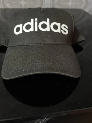 Svart keps från Adidas - Säljer en svart keps från Adidas med stor vit logga framtill. Klassisk böjd skärm och justerbar baksida. Tillverkad i slitstarkt bomullstyg, perfekt för en sportig och avslappnad stil.