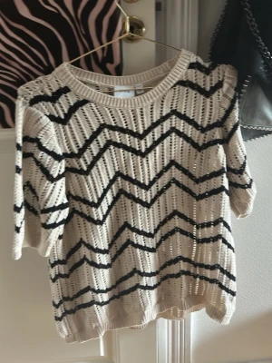 Beige stickad tröja från Vila - Säljer en beige stickad tröja från Vila med svarta sicksack-ränder. Tröjan har korta ärmar, rund halsringning och ett luftigt, hålmönstrat material som ger en avslappnad vibe. Perfekt att styla med jeans eller kjol för en trendig look.