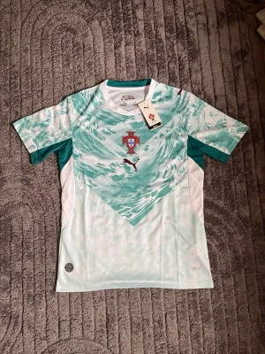 Portugal fotbollströja Puma grön/vit - Snygg Portugal fotbollströja från Puma i vitt och grönt med vågigt mönster. Tröjan har Portugal-emblemet på bröstet och Puma-logga i svart. Tillverkad i lätt och ventilerande material med DryCell-teknologi. Perfekt för träning eller match.