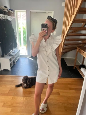 Vit kortklänning med volangkrage - Säljer en vit denim klänning med stora volanger och krage. Klänningen har knappar hela vägen framtill och är ärmlös. Perfekt för sommardagar och har en trendig, avslappnad passform. Finns en liten fläck!! 
