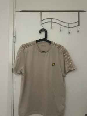 Beige t-shirt med loggan lyle & scott - Snygg beige t-shirt med korta ärmar och rund hals. Diskret logga i gult och svart på bröstet samt band med text längs axlarna. Perfekt till jeans eller shorts för en clean och stilren look.