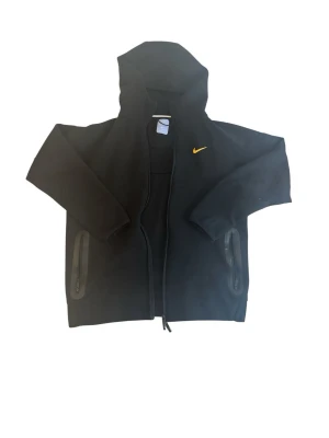 Svart Nike hoodie med dragkedja - Säljer en svart hoodie från Nike med gul broderad logga på bröstet. Hoodien har dragkedja framtill, två fickor med dragkedja och en stor huva. Materialet är mjukt och bekvämt, perfekt för chill dagar. Små broderade detaljer i gult på ryggen.