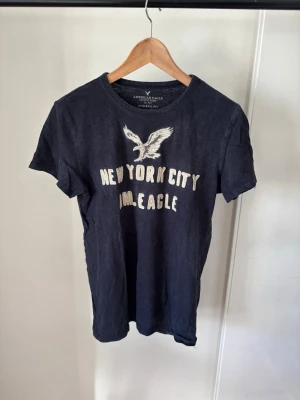 Svart t-shirt American Eagle NYC - Snygg svart t-shirt från American Eagle med tryck av en örn och texten 'New York City' och 'AM. EAGLE' på bröstet. Klassisk rund hals och korta ärmar. Perfekt för en avslappnad streetwear-look.
