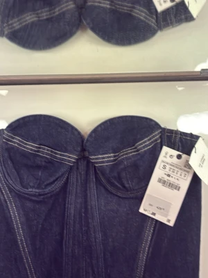 Mörkblå denim bustier tubtopp - Trendig mörkblå denim bustier tubtopp med markerade sömmar och formade kupor. Toppen är axelbandslös och har en croppad passform som ger en edgy look. Stretch och dragkedja i ryggen. Perfekt att styla med höga jeans eller kjol för en cool vibe "Även snygg under en vit skjorta för en mer sostifikerad lager på lager look eller en tunn, tajt polotröja. Det skapar en intressant silhuett och gör plagget mer användbart även under svalare dagar.