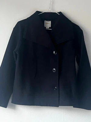Svart kort kappa med knappar  - Stilren svart kappa med stora slag och tre knappar framtill. Modellen är rak och något kortare, med långa ärmar och enkel design. Perfekt för dig som gillar minimalistisk och klassisk stil. Materialet ser ut att vara ullblandning.