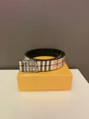 Burberry randigt bälte med B-spänne - Snyggt bälte från Burberry med klassiskt randigt mönster i beige, brunt och svart. Bältet har ett stort silvrigt B-spänne och är tillverkat i skinn. Perfekt accessoar för att lyfta din outfit.