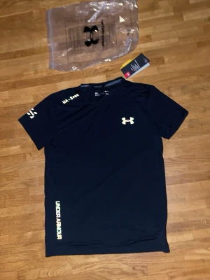 Svart tränings-tshirt från Under Armour - Svart kortärmad tränings-tshirt från Under Armour med logga på bröstet och tryck på ärm och nederkant. Tillverkad i lätt och snabbtorkande syntetmaterial, perfekt för träning. Modern passform och rund halsringning. storlek M men passar S