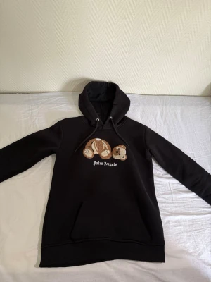 Svart Palm Angels hoodie med björn - Svart hoodie från Palm Angels med broderad nallebjörn på bröstet och vit logga under. Klassisk huva med dragsko och stor magficka framtill. Hoodien har ribbade muddar vid ärmslut och nederkant. Perfekt för dig som gillar streetwear och coola detaljer.
