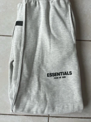 Grå Essentials Fear of God mjukisbyxor - Säljer ett par ljusgrå mjukisbyxor från Fear of God Essentials. Byxorna har resår i midjan och raka ben, samt svart logga tryckt på ena benet. Tillverkade i mjukt och bekvämt material, perfekta för en avslappnad stil. Skicka gärna frågor om det behövs och jag skickar gärna fler bilder. Jag hittar inte påsen och tagsen till byxorna fast jag ska försöka hitta de!