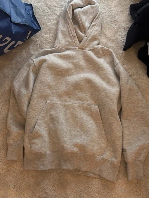 Ljusgrå hoodie med broderad detalj - Säljer en ljusgrå hoodie med huva och stor magficka. Tröjan har ribbade muddar vid ärmar och nederkant samt en diskret broderad logga på ena ärmen. Tillverkad i mjukt sweatshirtmaterial, perfekt för chill dagar.