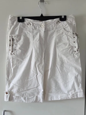Vita boyshorts/capris med spetsdetaljer - Säljer ett par vita boyshorys/capri byxor med snygga spets och nitdetaljer. Shortsen har knappstängning, bälteshällor och dekorativa knappar vid benslut. Passar perfekt till sommaren. Storleken är L men lite små i storlek så passar mer som en M.