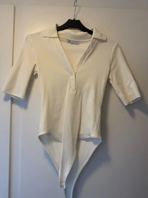 Vit body med krage från Zara - Stilren vit body från Zara med klassisk krage och trekvartsärm. Modellen har knäppning framtill med tre knappar och mjukt, stretchigt material som sitter snyggt på kroppen. Perfekt till både jeans och kjol för en clean look.