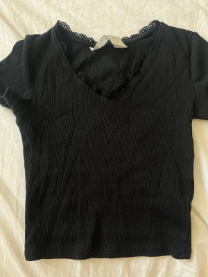Svart ribbad topp med spetsdetalj - Snygg svart ribbad topp med korta ärmar och v-ringning. Spetsdetaljer längs halsringningen ger en extra touch. Perfekt att styla till jeans eller kjol för en trendig look.