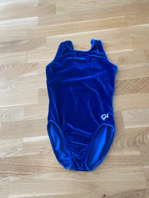 Blå sammetsgymnastikdräkt från GK - Snygg blå gymnastikdräkt i sammetsmaterial från GK. Dräkten har klassisk skärning med breda axelband och en liten vit GK-logga broderad framtill. Perfekt för träning eller tävling inom gymnastik. Den är till Järfälla gymnasterna