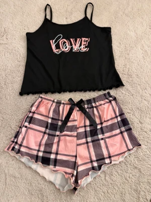 Pyjamasset - Sött pyjamasset med svart linne med tunna axelband och LOVE-tryck i rosa och vitt. Linnet har vågig kant nedtill. Tillhörande shorts i rosa med svartvitt rutmönster och svart rosett i midjan. Perfekt för mysiga kvällar. - helt oanvänd 
