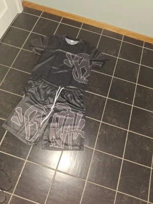 Syna world x central cee svart - Cool svart t-shirt med stora vita graffiti-inspirerade tryck på både fram- och baksidan. T-shirten har rund hals och korta ärmar. Materialet känns som syntet, vilket ger en sportig vibe. Perfekt för dig som gillar streetwear och vill sticka ut.