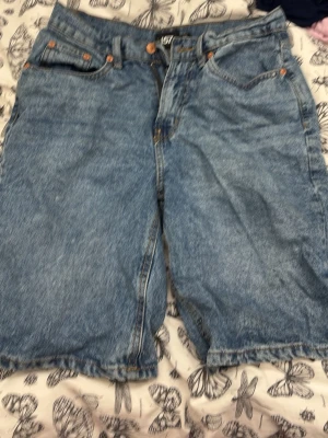 Blå jeansshorts i baggy modell. Jorts - Säljer ett par klassiska blå jeansshorts i baggy modell med fem fickor och knappgylf. Shortsen har en avslappnad passform och är tillverkade i slitstarkt denimtyg. Perfekta för en chill och avslappnad stil. Jorts