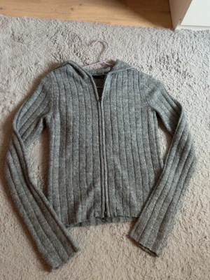 Grå ribbstickad hoodie med dragkedja - Mysig grå ribbstickad hoodie med dragkedja framtill och långa ärmar. Tröjan har huva och tumhål i ärmsluten för extra komfort. Perfekt för lager på lager och chill dagar. Materialet känns mjukt och stretchigt.