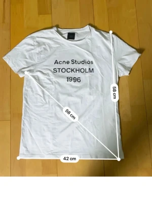 Vit Acne Studios t-shirt Stockholm - Vit t-shirt från Acne Studios med trycket 'Acne Studios STOCKHOLM 1996' på bröstet. Klassisk passform med rund hals och korta ärmar. Perfekt basic-plagg för en clean och stilren look.