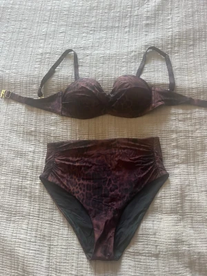Leopardmönstrad bikini i mörkbrun - Snygg bikini med leopardmönster i mörkbruna toner. Överdelen har bygel och reglerbara axelband, medan underdelen är högmidjad för en trendig look. Materialet känns mjukt och stretchigt, perfekt för strandhäng.