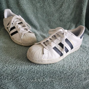 Vita Adidas Superstar sneakers - Klassiska vita Adidas Superstar sneakers med svarta ränder på sidorna och den ikoniska snäckformade tån. Skorna har vita skosnören och en låg silhuett. Perfekta för dig som gillar retrostil och streetwear.
