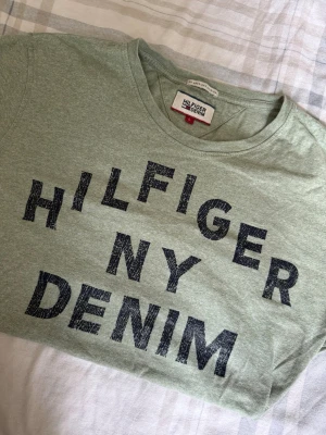 Omsydd Grön Hilfiger Denim t-shirt - Snygg ljusgrön t-shirt från Hilfiger Denim med stort tryck i mörkblått framtill. Klassisk rund halsringning och korta ärmar. Perfekt för en avslappnad och trendig look. Tillverkad i mjuk bomull för skön komfort.