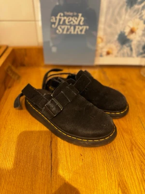 Svarta mockasandaler dr martens  - Säljer ett par svarta sandaler i mocka med justerbart spänne och öppen häl. Sulan är robust med gul kontrastsöm runt om, vilket ger en cool och trendig look. Perfekta för dig som vill ha något bekvämt men ändå stilrent till sommaren.