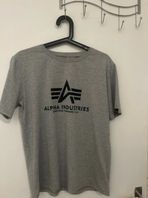 Grå Alpha Industries t-shirt - Snygg grå t-shirt från Alpha Industries med svart logga och text på bröstet. Klassisk rund halsringning och korta ärmar. Perfekt till jeans eller shorts för en avslappnad look.