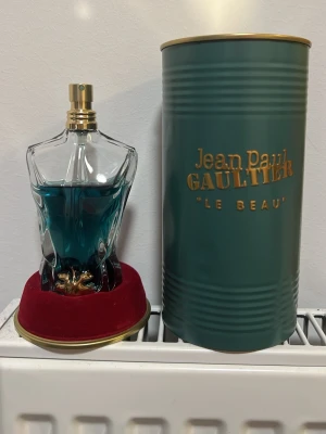 Jean Paul Gaultier Le Beau parfym - Säljer en Jean Paul Gaultier Le Beau parfym i en unik flaska formad som en manlig torso med blågrön vätska. Flaskan har gulddetaljer och kommer med en elegant grön metalltub med guldfärgad text. Basen är klädd i röd sammet med guldkant.