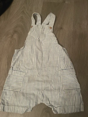 Randiga hängselshorts i bomull - Säljer ett par ljusblå och vita randiga hängselshorts med breda axelband och två stora fickor framtill. Shortsen har klassisk design och är tillverkade i mjuk bomull, perfekta för en avslappnad stil. För barn 18-24 månader