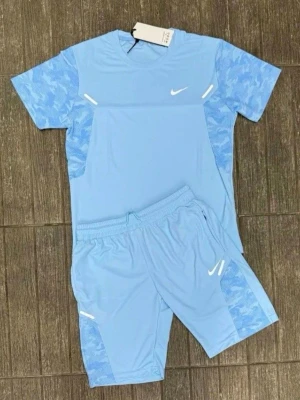 Ljusblått träningsset från Nike - Säljer ett ljusblått träningsset från Nike med t-shirt och shorts. Setet har mönstrade detaljer på ärmar och sidor, samt vita Nike-loggor på både tröja och shorts. Perfekt för träning eller sportaktiviteter.