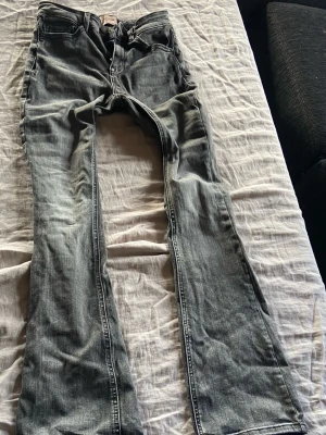 Grå bootcut jeans med slitningar - Säljer ett par grå bootcut jeans med snygga slitningar och tvättad look. Jeansen har klassiska fem fickor och dragkedja med knapp. Perfekta för dig som gillar en avslappnad men trendig stil.