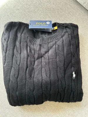 Svart kabelstickad tröja Polo Ralph Lauren - Kabelstickad svart tröja från Polo Ralph Lauren med rund halsringning och den klassiska vita logobroderingen på bröstet. Tröjan har långa ärmar och ribbade muddar vid ärmslut och nederkant. Perfekt för en stilren och tidlös look.               PRISET KAN DISKUTERAS 