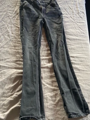 Grå bootcut jeans med slitningar - Säljer ett par grå bootcut jeans med lätt slitna detaljer och klassiska fickor både fram och bak. Jeansen har en något utsvängd passform nertill och är tillverkade i denim med synliga sömmar. Perfekta för en avslappnad och trendig look.