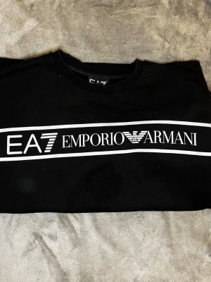Svart sweatshirt från Emporio Armani - Säljer en Snygg svart sweatshirt från Emporio Armani EA7 med vit logga och text över bröstet. Tröjan har rund halsringning, långa ärmar och ribbade muddar vid ärmslut och nederkant. Perfekt för en trendig och avslappnad stil. Vid frågor hör av er priset kan alltid diskuteras 
