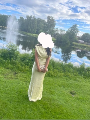 Ljusgul långklänning med oneshoulder - Elegant ljusgul långklänning i satinliknande material med oneshoulder-design och draperad överdel. Klänningen har en figurnära passform och slät yta som ger en lyxig känsla. Perfekt för speciella tillfällen. Använd en gång. 