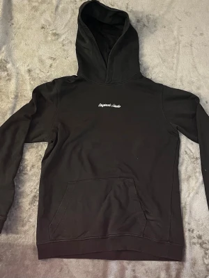 Mörkgrön  hoodie från Originals Studio - Säljer en Snygg mörkgrön hoodie från Jack&jones med broderad vit text på bröstet. Klassisk känguruficka framtill och stor huva. Tillverkad i mjukt bomullsmaterial som känns skönt mot huden. Perfekt för en avslappnad och trendig look. Vid frågor hör av er priset kan alltid diskuteras 