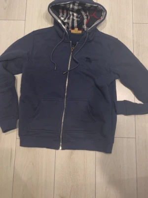 Marinblå hoodie från Burberry - Snygg marinblå hoodie från Burberry med dragkedja, fickor framtill och klassiskt rutigt foder i huvan. Broderad Burberry-logga på bröstet och justerbar dragsko i huvan. Perfekt för en avslappnad och stilren look.