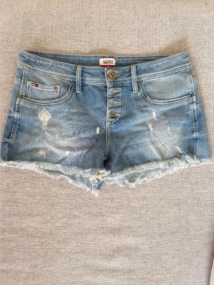 Blå jeansshorts från Tommy Hilfiger stl31 - Ett par denimshorts från Hilfiger Denim med en ljus indigofärg och ”destructed” detaljer. Shortsen har en knappgylf och fransiga benslut. Fickor finns fram och bak. Midjemått 42cm Längd 28cm Helt nya oanvända! Djur och rökfritt hem.