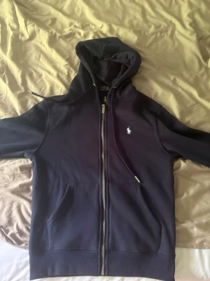 Mörkblå hoodie från Polo Ralph Lauren - Snygg mörkblå hoodie med dragkedja. Klassisk design med vit broderad logga på bröstet, två fickor framtill och justerbar huva med snören. Tillverkad i mjukt bomullsmaterial som är skönt att ha på sig. Storlek s-xs 