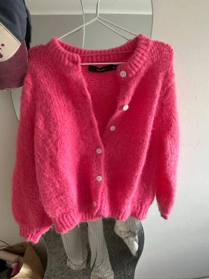 Rosa fluffig kofta med knappar - Supermjuk och fluffig rosa kofta med vita knappar framtill. Koftan har en oversized passform, ribbade muddar och rund halsringning. Perfekt för dig som vill ha en mysig och trendig look. Materialet känns som en mohairmix.