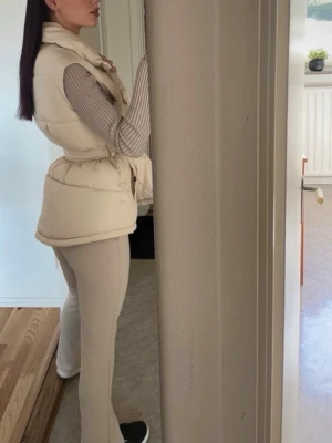Beige dunväst med knytskärp - Stilren beige dunväst med bred krage och knytskärp i midjan. Västen har en puffig, quiltad design och ger en trendig siluett. Perfekt att bära över en långärmad tröja för en snygg lager-på-lager-look. Inga synliga loggor eller märken.