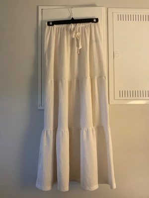 Vit långkjol med volanger och knyt - Säljer en vit långkjol med volangdetaljer och resår i midjan samt knytband. Kjolen har en luftig och flowy känsla, perfekt för varma dagar. Materialet är mjukt och har en lätt struktur som ger extra dimension till looken.