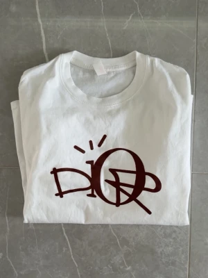 Vit t-shirt med brun DIOR-grafik - Vit t-shirt från Dior med stor brun grafisk logga på bröstet. Klassisk rund hals och korta ärmar. Tillverkad i mjuk bomull för en bekväm känsla och enkel stil.