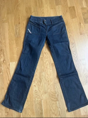 Mörkblå bootcut jeans från Diesel - Snygga mörkblå jeans från Replay med bootcut passform och raka ben. Jeansen har två stora framfickor och en liten Replay-logga på vänster ficka. Klassisk denimlook med bälteshällor och knappgylf. Perfekta för en avslappnad och trendig stil.