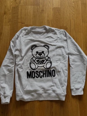 Vit Moschino sweatshirt med björn - Säljer en vit sweatshirt från Moschino med det ikoniska Toy-björnmotivet och stor svart logga framtill. Tröjan är i 100% bomull och har rund hals samt ribbade muddar vid ärmar och nederkant. Perfekt för dig som gillar streetwear och statement-plagg.