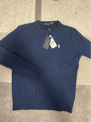 Polo ralph lauren sweater tröja - Mörkblå kabelstickad tröja från Polo Ralph Lauren med rund halsringning och den klassiska broderade loggan i vitt på bröstet. Tröjan har långa ärmar och ribbade muddar. Materialet är mjukt och stretchigt, perfekt för en snygg och avslappnad look.