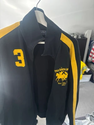 Svart gul  hoodie från Polo Ralph Lauren - Cool svart gul hoodie från Polo Ralph Lauren med gula detaljer och broderad logga på bröstet. Dragkedja framtill och hög krage. Inte använt på länge. Storlek L men passar mer M
