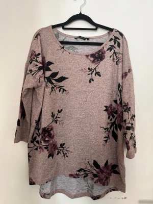 Ljusrosa blommig långärmad tröja - En ljusrosa långärmad tröja med stora blommönster i mörklila och svart. Tröjan har en rund halsringning och är lite längre i modellen.
